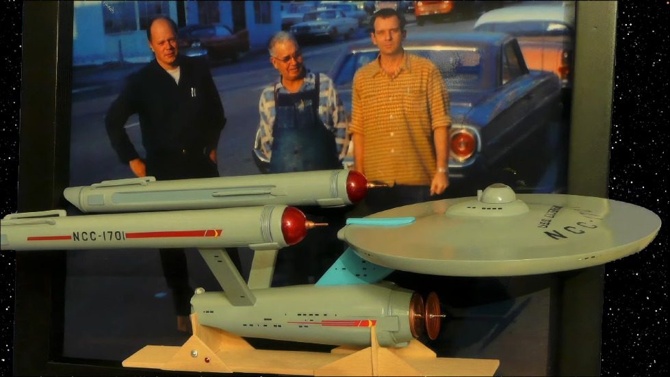 A group of men standing next to a model of a space ship Description automatically generated