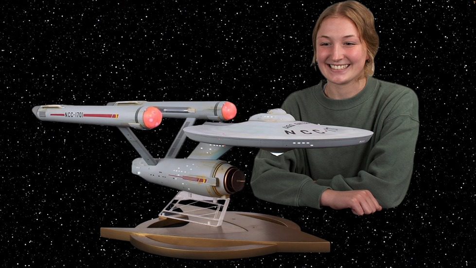 A person smiling next to a model of a space ship Description automatically generated